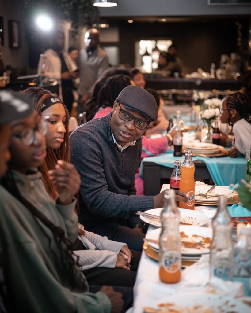 A man holding a bottle of fanta with a group of people over Nigerian Dishes.